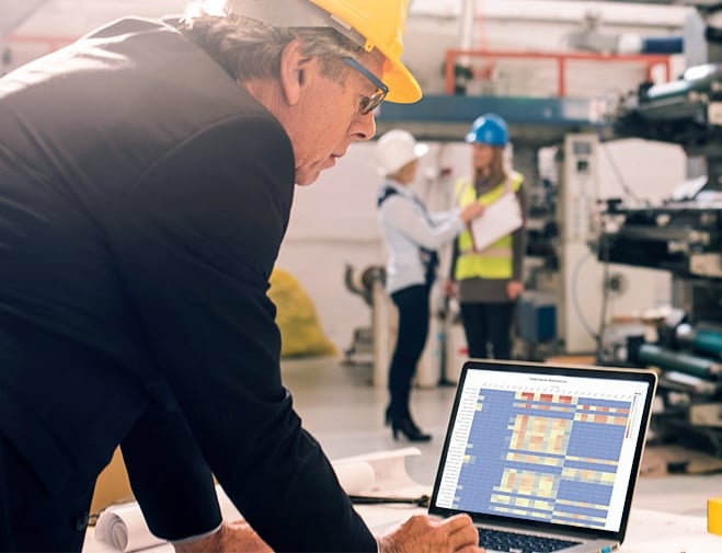 "Manufacturing manager in a hard hat reviewing a heat map dashboard on a laptop in a factory setting. Three other engineers in safety gear discuss a report in the background. The image represents industrial data analysis, production efficiency, and smart manufacturing technology."